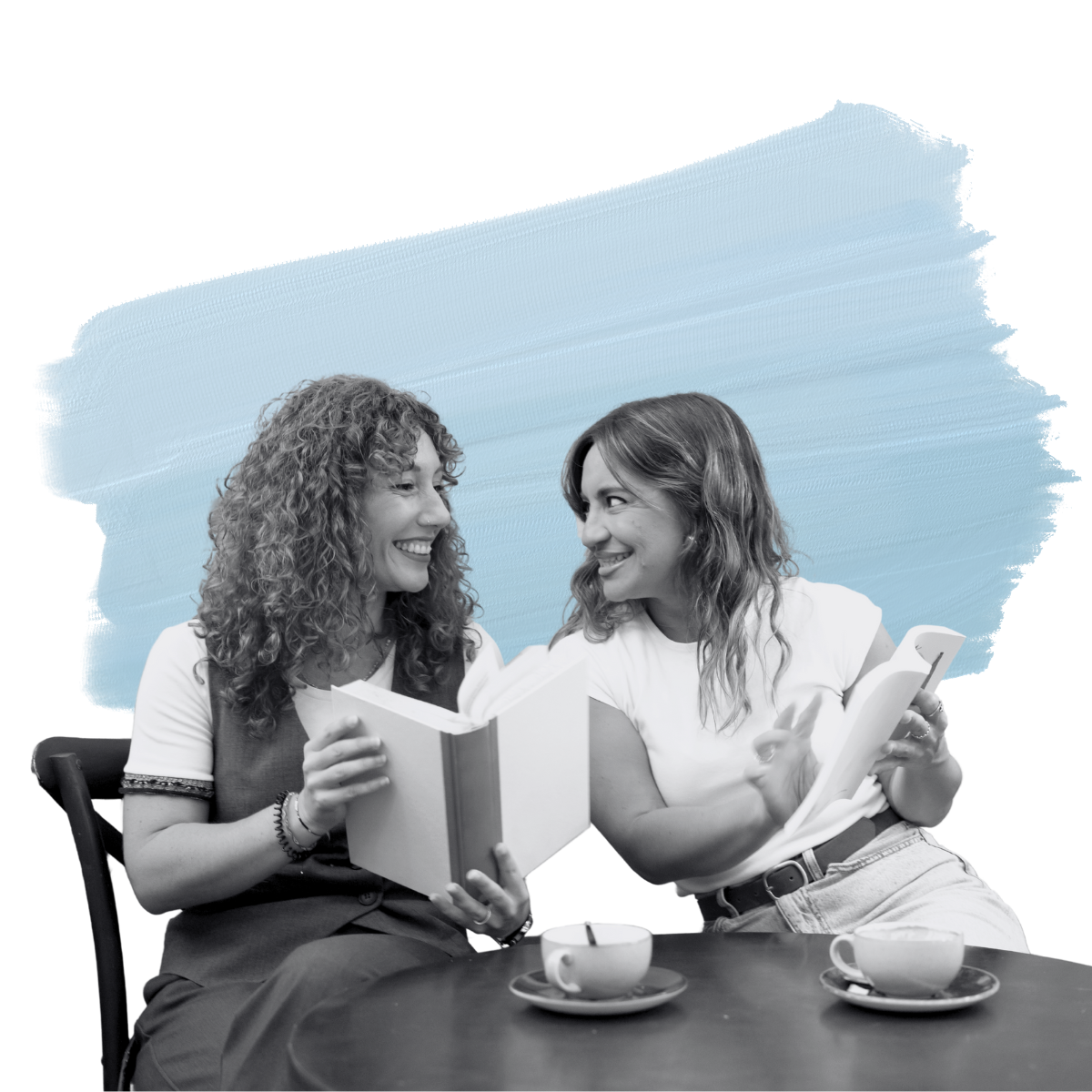 Two women sitting at a coffee table holding books and smiling at eachother